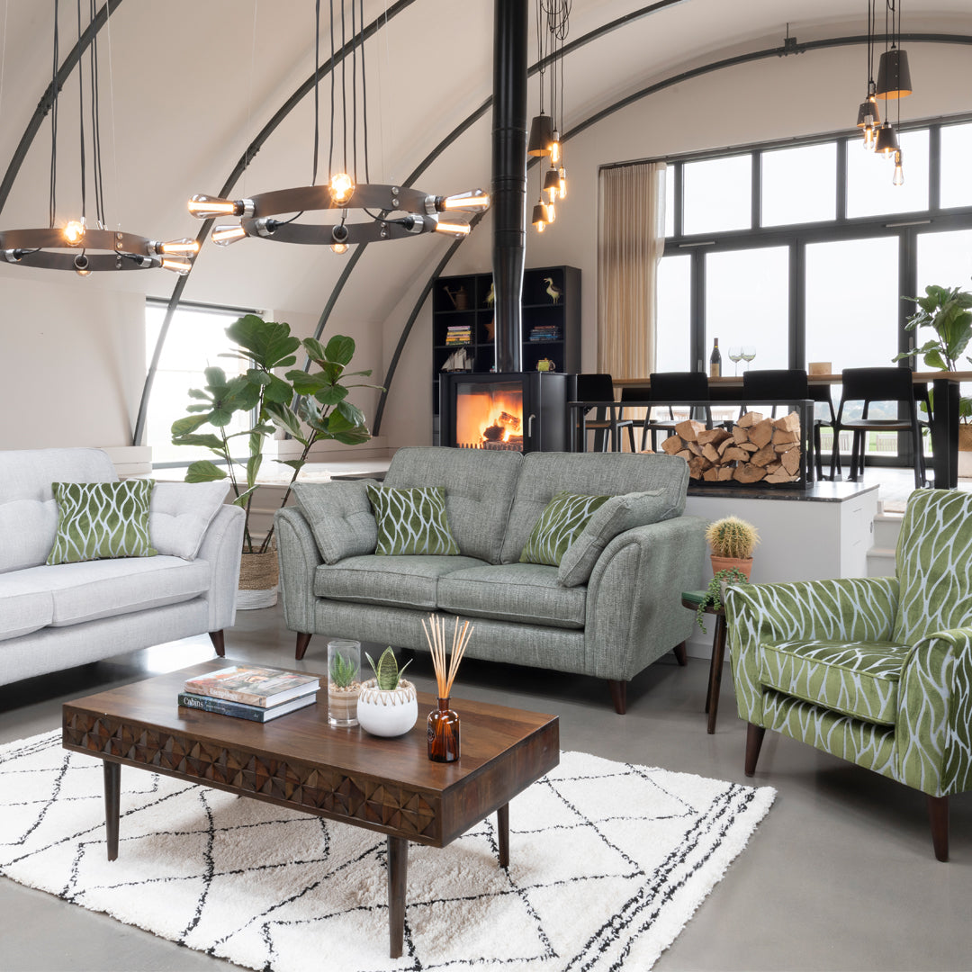 This image show's a grey sofa with green scatter cushions which match the green accent chairs. The sofa is set in a cosy living room with a roaring fire and moody lighting. 