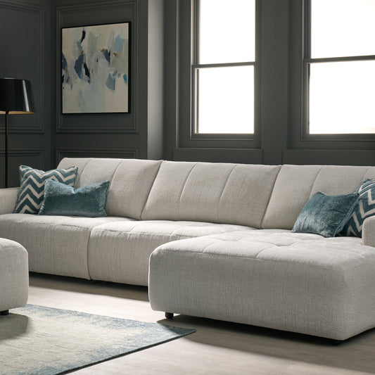 A beige Lucca Chaise Lounge sofa with blue accent pillows, positioned in a room with a large window and decorative wall art.
