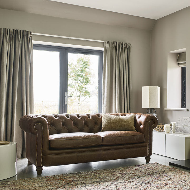A lifestyle image of the Abraham Junior in a brown leather. The sofa is against beige pleated curtains and a beige rug. 