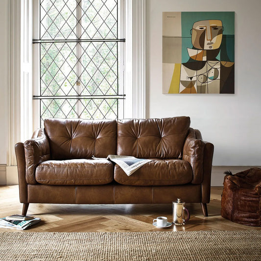 A luxurious brown leather sofa with squishy arm cushions, placed in a well-lit living room with a modern abstract painting on the wall, next to a wooden side table with a lamp and a magazine.