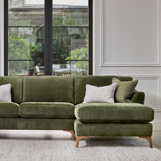 A green Jasmin Chaise Sofa with supportive back cushions and a sleek design, with foot shown in image: Smoke Wood, placed in a room with large windows.