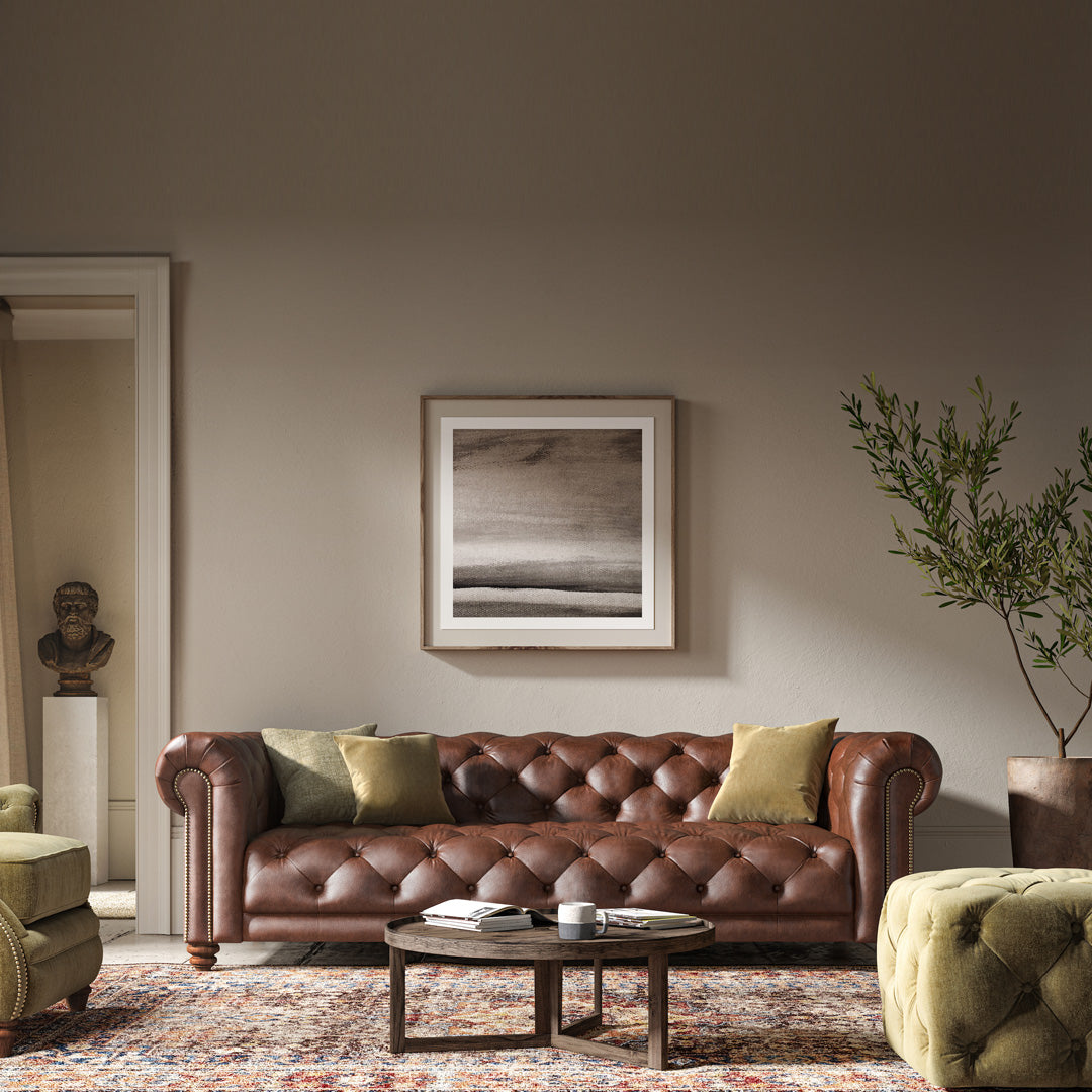 Elegant traditional Chesterfield style sofa with deep seat and tufts, thick scroll arms with studding, and low bulbous wooden feet, placed in a well-lit living room with a neutral wall background, a framed abstract picture above it, a green armchair to the side, and a round brown ottoman in front.