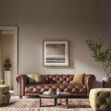 Elegant traditional Chesterfield style sofa with deep seat and tufts, thick scroll arms with studding, and low bulbous wooden feet, placed in a well-lit living room with a neutral wall background, a framed abstract picture above it, a green armchair to the side, and a round brown ottoman in front.