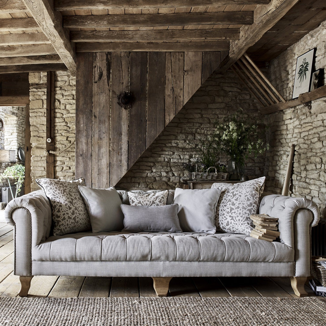 A classic chesterfield design sofa with deep buttoned seats and thickly pleated roll arms, placed in a rustic living room setting with wooden beams on the ceiling and a stone wall.