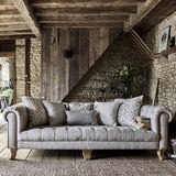 A classic chesterfield design sofa with deep buttoned seats and thickly pleated roll arms, placed in a rustic living room setting with wooden beams on the ceiling and a stone wall.