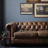 A lifetsyle image of the Abraham Junior Grand Sofa in bornw leather. The sofa is against a blue wall and sitting next to the sofa is a glass vase with flowers. 