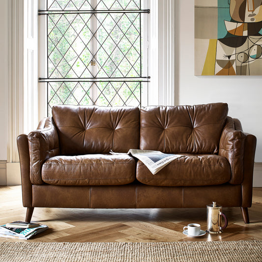 A lifestyle image of the Saddler Armchair in brown. The sofa has a newspaper in the middle and a cafietiere on the floor by the right side of it. 