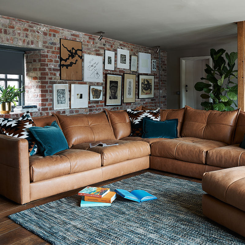 A lifestyle image of the Alexander and James Tod conrer Sofa. The tod is shown against a brick wall and blue plush cushions scattered along the sofa.