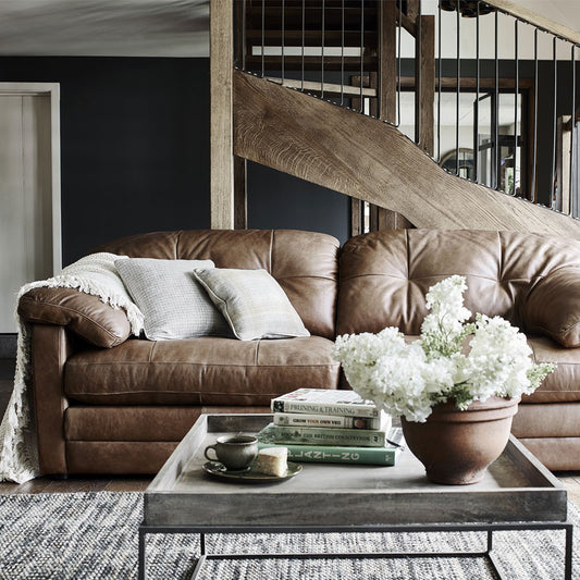 A lifestyle image of the Alexander & James Bailey 2 Seater Sofa. The sofa has cream cushions and a cream blanket styled on the left hand side of the sofa. The sofa sits against a wooden beam staircase . 