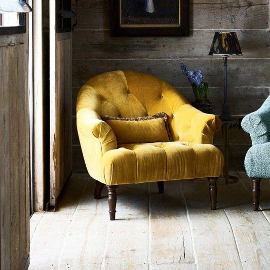 An lifestyle image of the Alexander & James Imogen Buttoned Chair.  The chair is in a yellow fabric with dark brown legs. The chair is positioned in front of a wooden panelled wall and a thin lampshade next to it. 